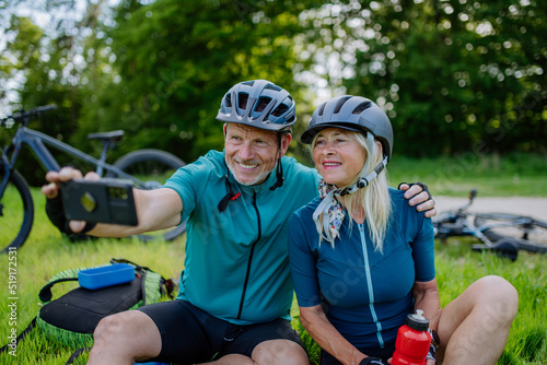 Active senior couple resting after bicycle ride at summer park, sitting on grass and taking slefie.