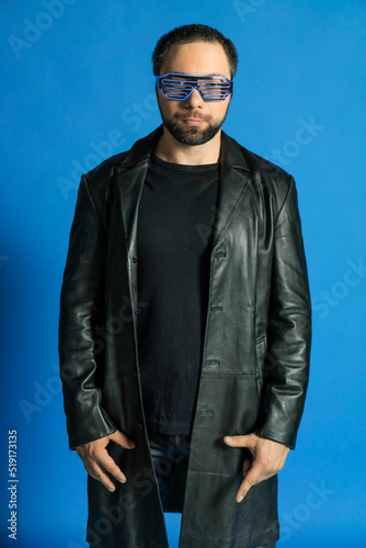 portrait of a man in a studio in a black leather coat. model with a beard on a blue background. trendy luminous glasses on the face