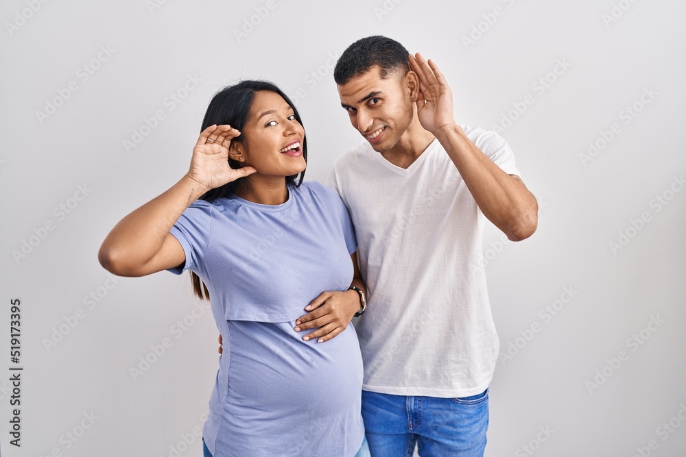 Young hispanic couple expecting a baby standing over background smiling with hand over ear listening an hearing to rumor or gossip. deafness concept. Young hispanic couple expecting a baby standing over background smiling with hand over ear listening an hearing to rumor or gossip. deafness concept.