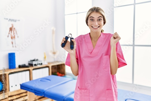 Young blonde woman working at pain recovery clinic holding hand strengthener screaming proud  celebrating victory and success very excited with raised arm