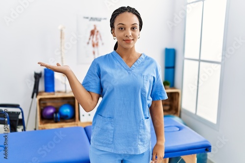 Young african american woman working at pain recovery clinic smiling cheerful presenting and pointing with palm of hand looking at the camera.