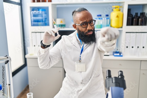 African american man working at scientist laboratory holding syringe pointing with finger to the camera and to you  confident gesture looking serious