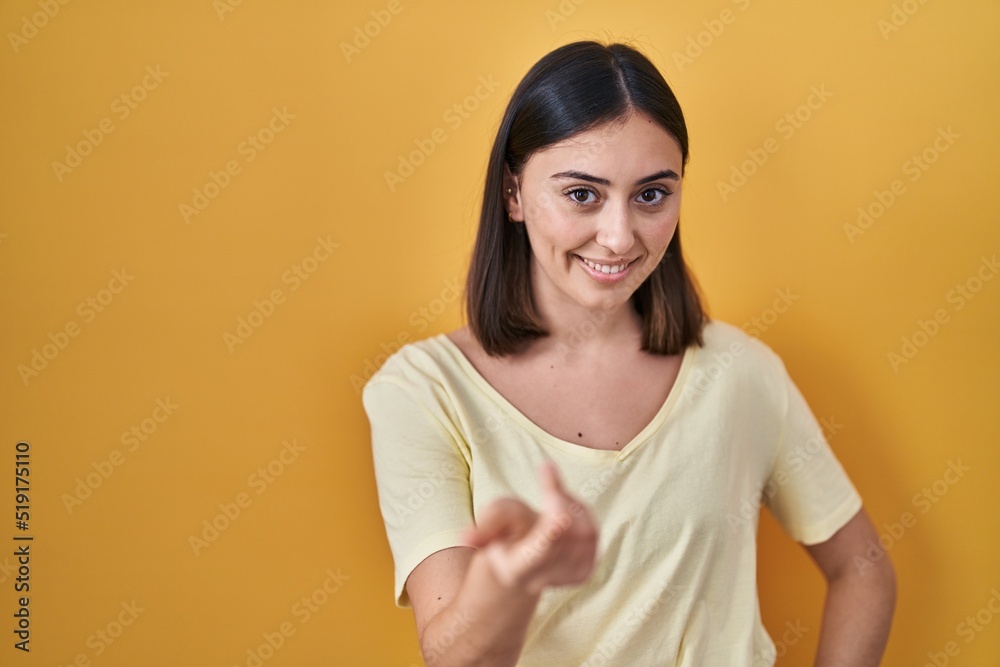 Obraz premium Hispanic girl wearing casual t shirt over yellow background beckoning come here gesture with hand inviting welcoming happy and smiling