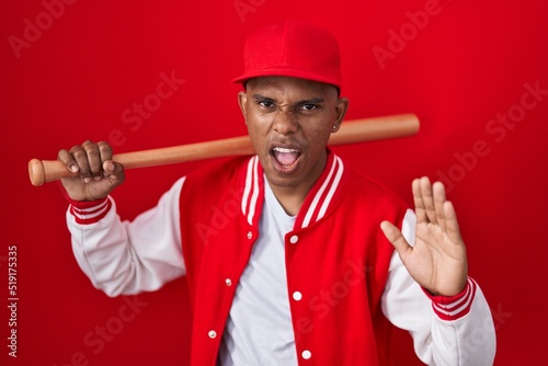 Young hispanic man playing baseball holding bat angry and mad screaming frustrated and furious, shouting with anger. rage and aggressive concept.