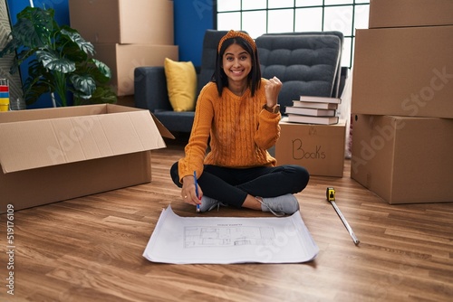 Young hispanic woman sitting on the floor at new home looking at blueprints screaming proud, celebrating victory and success very excited with raised arms photo