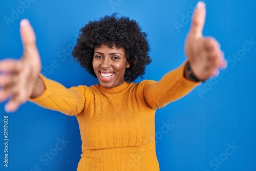 Black woman with curly hair standing over blue background looking at the camera smiling with open arms for hug. cheerful expression embracing happiness.