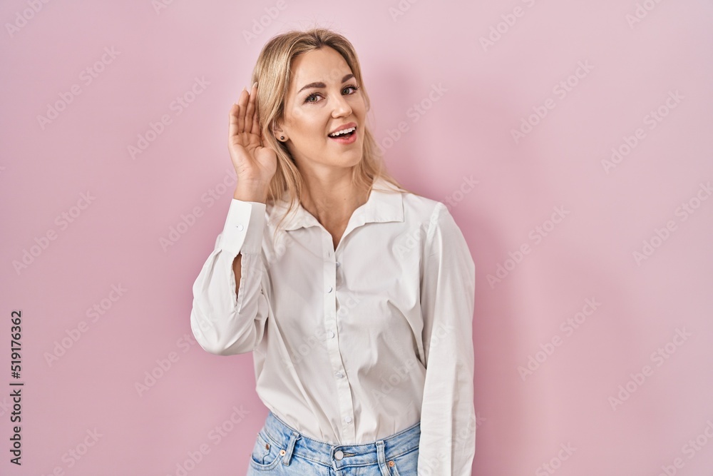 Young caucasian woman wearing casual white shirt over pink background smiling with hand over ear listening an hearing to rumor or gossip. deafness concept. Young caucasian woman wearing casual white shirt over pink background smiling with hand over ear listening an hearing to rumor or gossip. deafness concept.