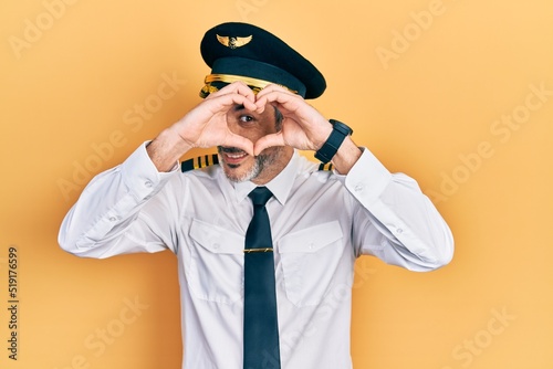 Handsome middle age man with grey hair wearing airplane pilot uniform doing heart shape with hand and fingers smiling looking through sign