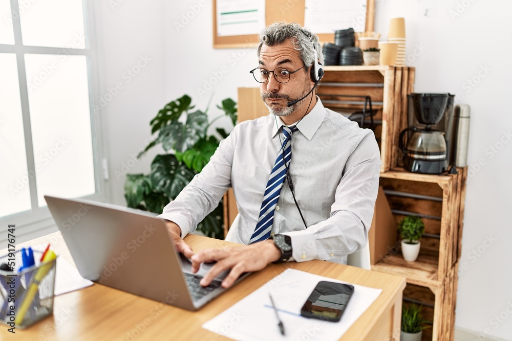 Middle age hispanic business man working at the office wearing operator headset puffing cheeks with funny face. mouth inflated with air, crazy expression. Middle age hispanic business man working at the office wearing operator headset puffing cheeks with funny face. mouth inflated with air, crazy expression.