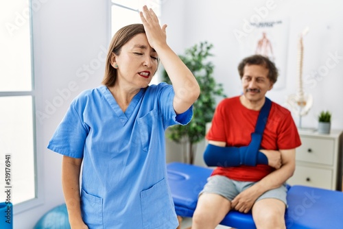 Middle age doctor woman with patient with arm injury at rehabilitation clinic surprised with hand on head for mistake  remember error. forgot  bad memory concept.