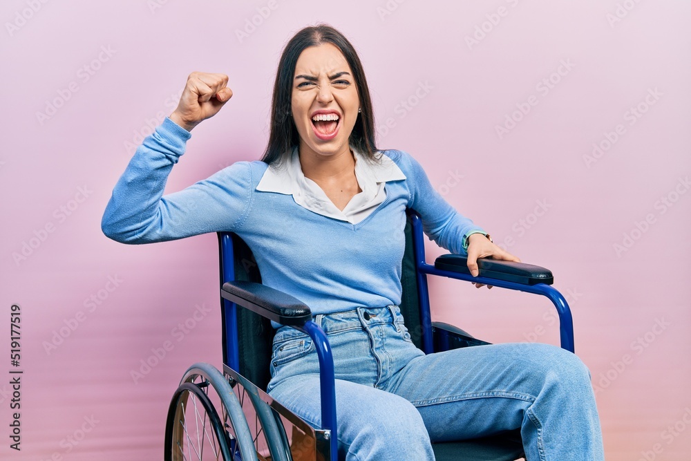 Beautiful woman with blue eyes sitting on wheelchair angry and mad raising fist frustrated and furious while shouting with anger. rage and aggressive concept.