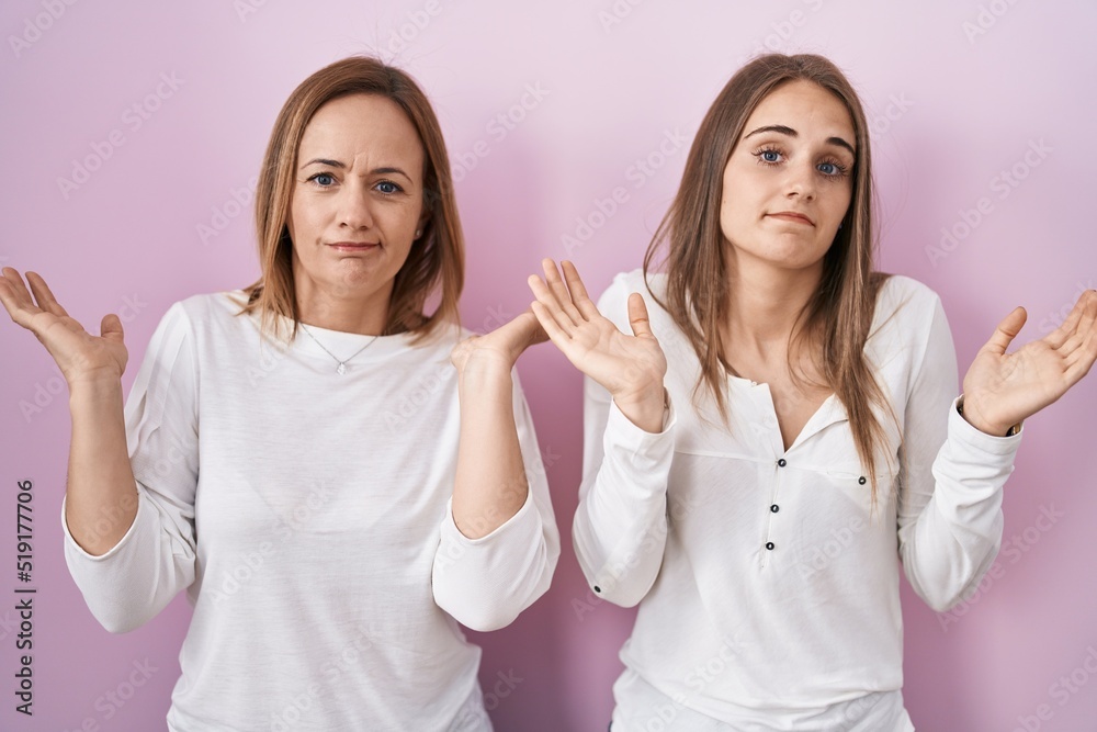 Middle age mother and young daughter standing over pink background clueless and confused expression with arms and hands raised. doubt concept. Middle age mother and young daughter standing over pink background clueless and confused expression with arms and hands raised. doubt concept.