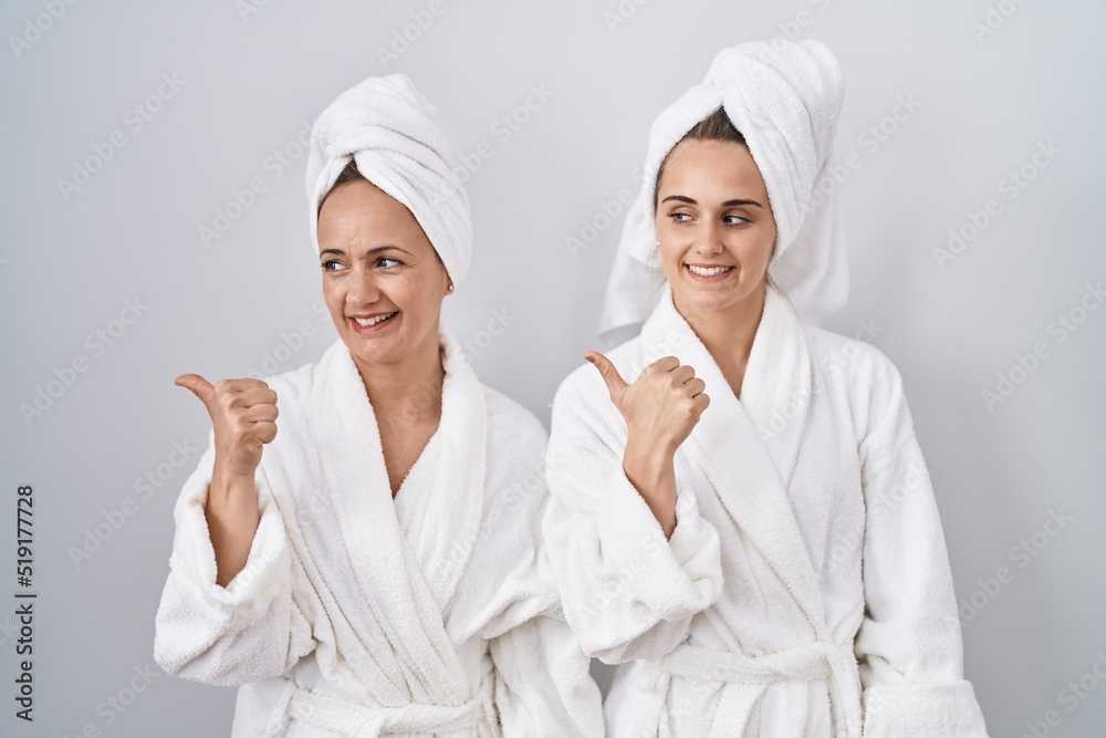 Middle age woman and daughter wearing white bathrobe and towel smiling with happy face looking and pointing to the side with thumb up.