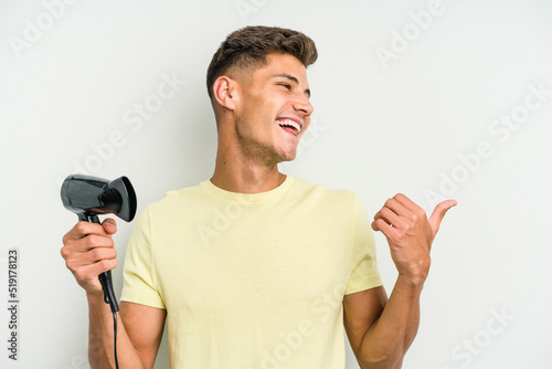 Young caucasian man holding hairdryer isolated on white background points with thumb finger away, laughing and carefree.