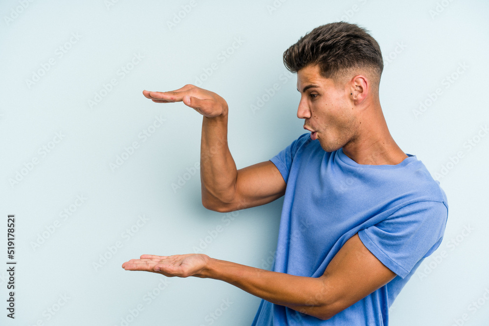 Young caucasian man isolated on blue background shocked and amazed holding a copy space between hands. Young caucasian man isolated on blue background shocked and amazed holding a copy space between hands.