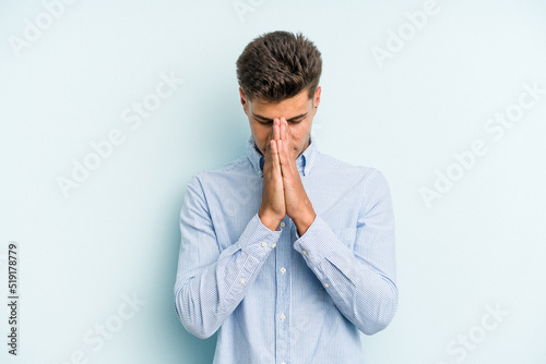Young caucasian man isolated on blue background praying, showing devotion, religious person looking for divine inspiration.