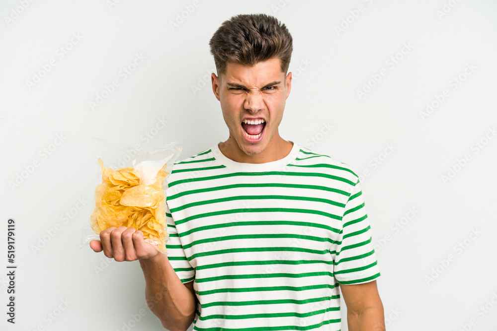 Young caucasian man holding crisps isolated on white background screaming very angry and aggressive. Young caucasian man holding crisps isolated on white background screaming very angry and aggressive.