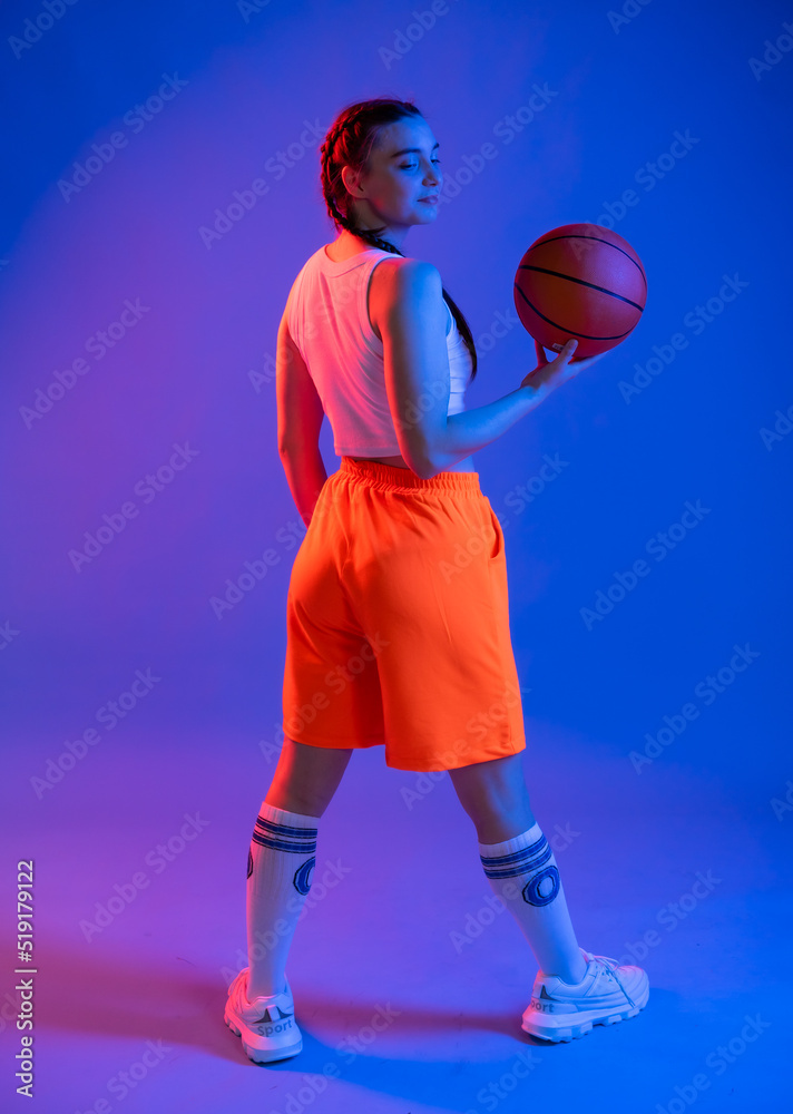 Naklejka premium a girl with a basketball in orange shorts, a t-shirt and a baseball cap. sportswoman in golfs and sneakers on a blue background. Physical education teacher. basketball player