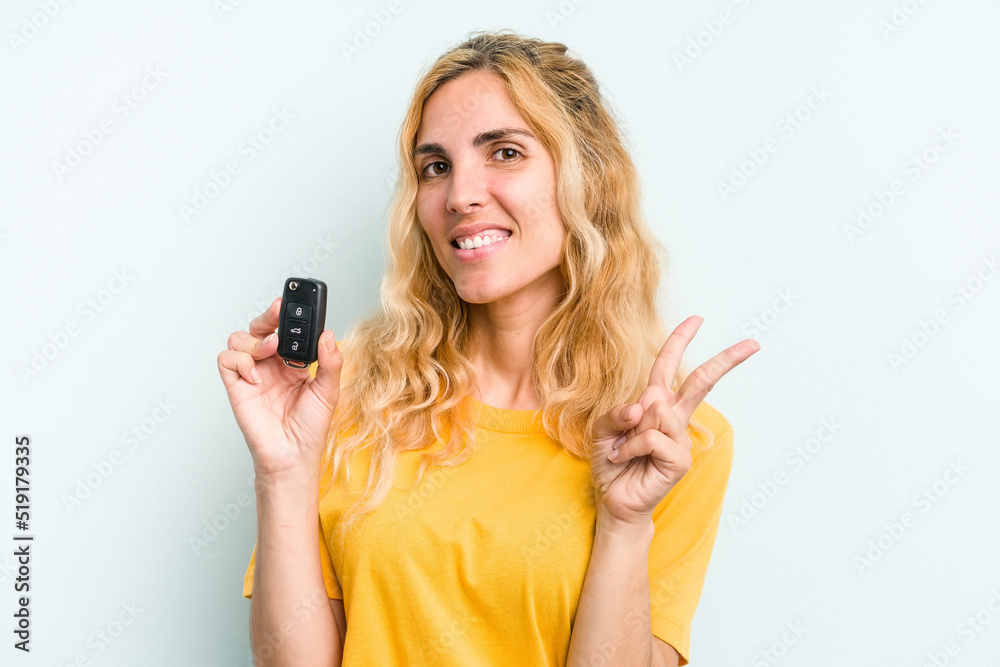 Young caucasian woman holding car keys isolated on blue background joyful and carefree showing a peace symbol with fingers. Young caucasian woman holding car keys isolated on blue background joyful and carefree showing a peace symbol with fingers.