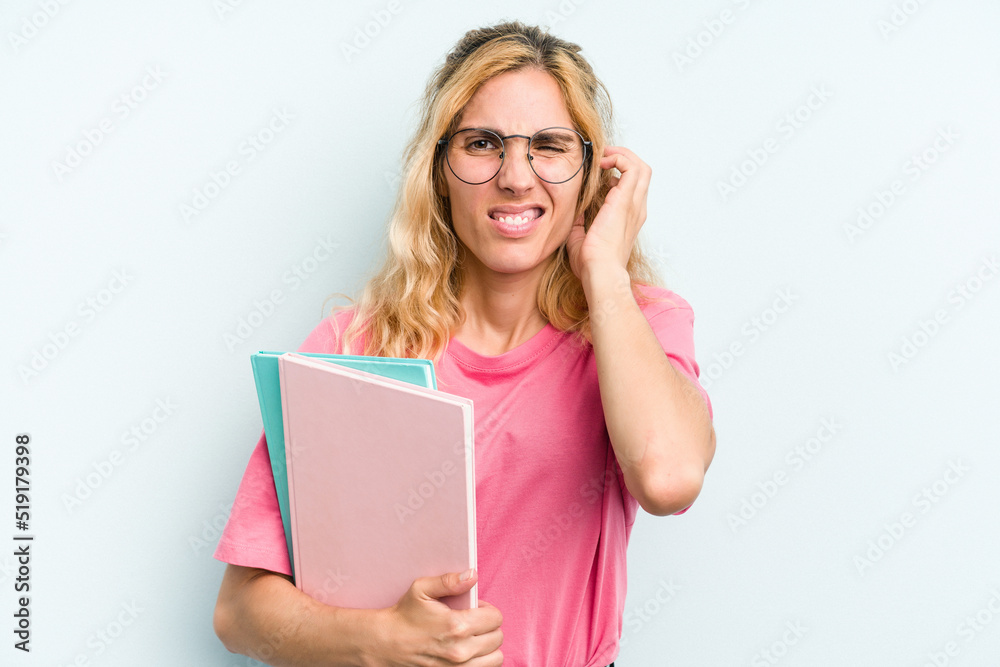 Young student caucasian woman holding books isolated on blue background covering ears with hands. Young student caucasian woman holding books isolated on blue background covering ears with hands.