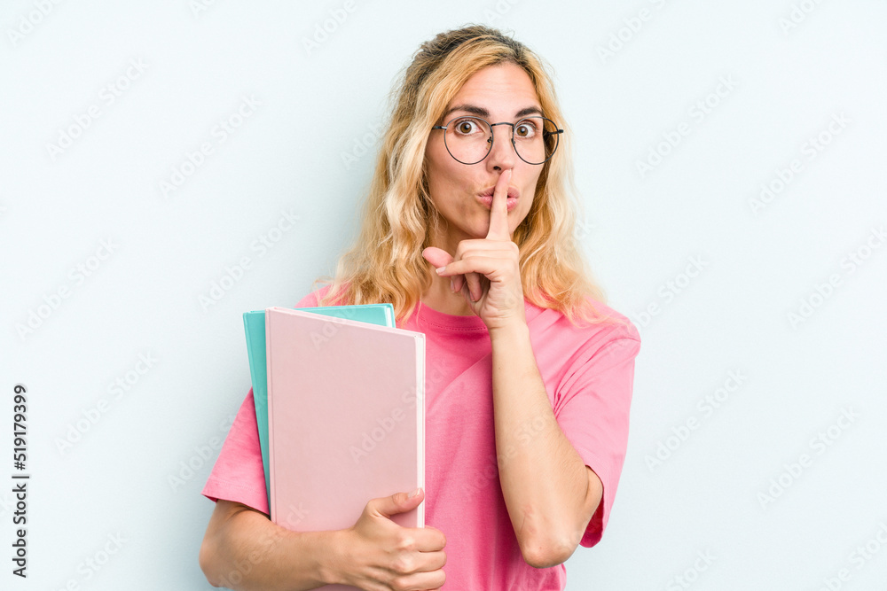 Young student caucasian woman holding books isolated on blue background keeping a secret or asking for silence. Young student caucasian woman holding books isolated on blue background keeping a secret or asking for silence.