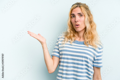 Young caucasian woman isolated on blue background impressed holding copy space on palm.