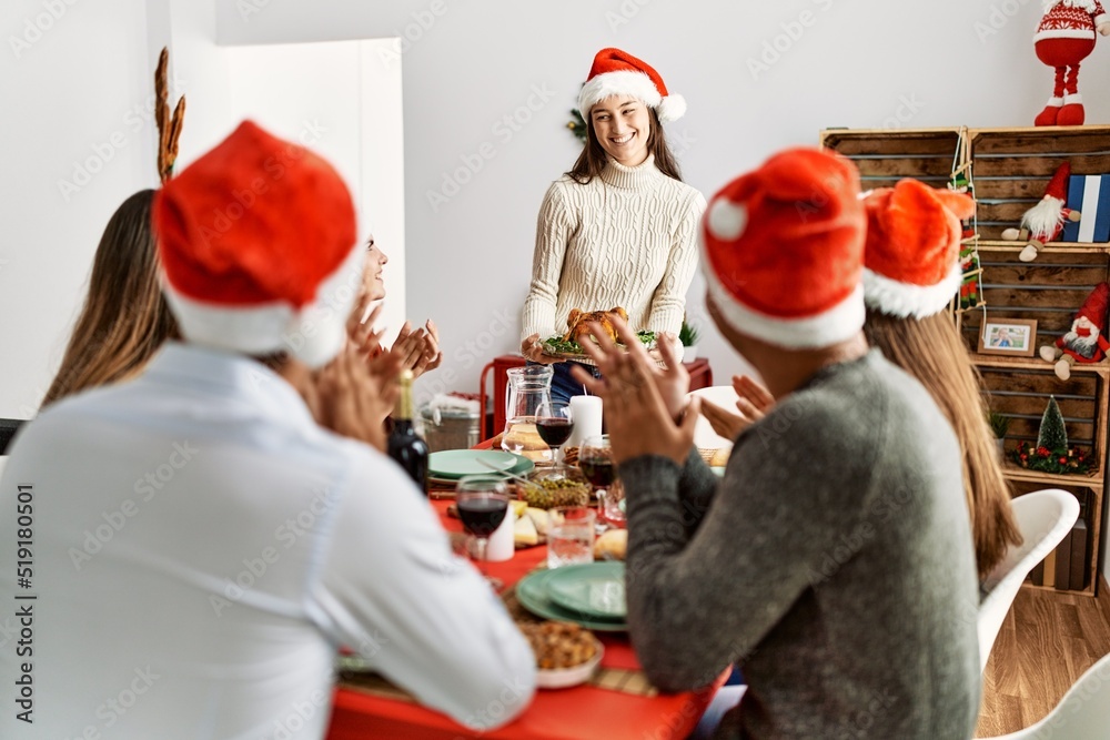 Fototapeta premium Group of people meeting clapping and sitting on the table. Woman standing and holding roasted turkey celebrating Christmas at home
