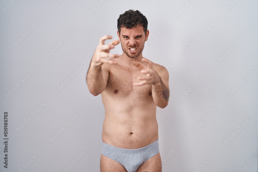 Young hispanic man standing shirtless wearing underware shouting frustrated with rage, hands trying to strangle, yelling mad
