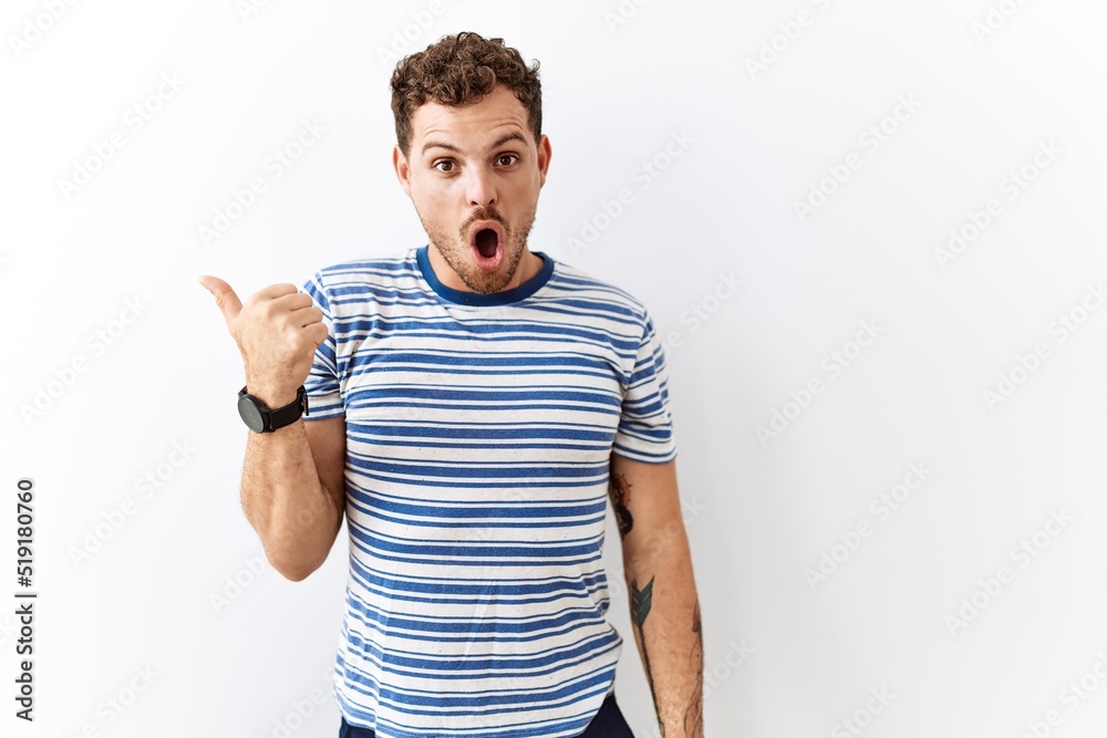 Handsome young man standing over isolated background surprised pointing with hand finger to the side, open mouth amazed expression. Handsome young man standing over isolated background surprised pointing with hand finger to the side, open mouth amazed expression.