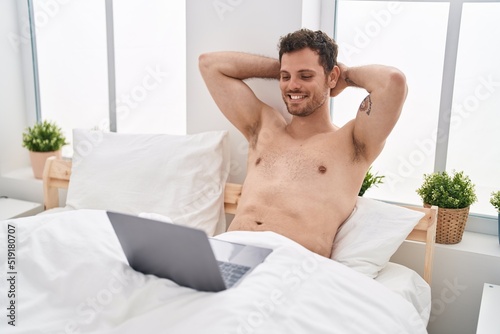 Young hispanic man using laptop relaxed with hands on head at bedroom