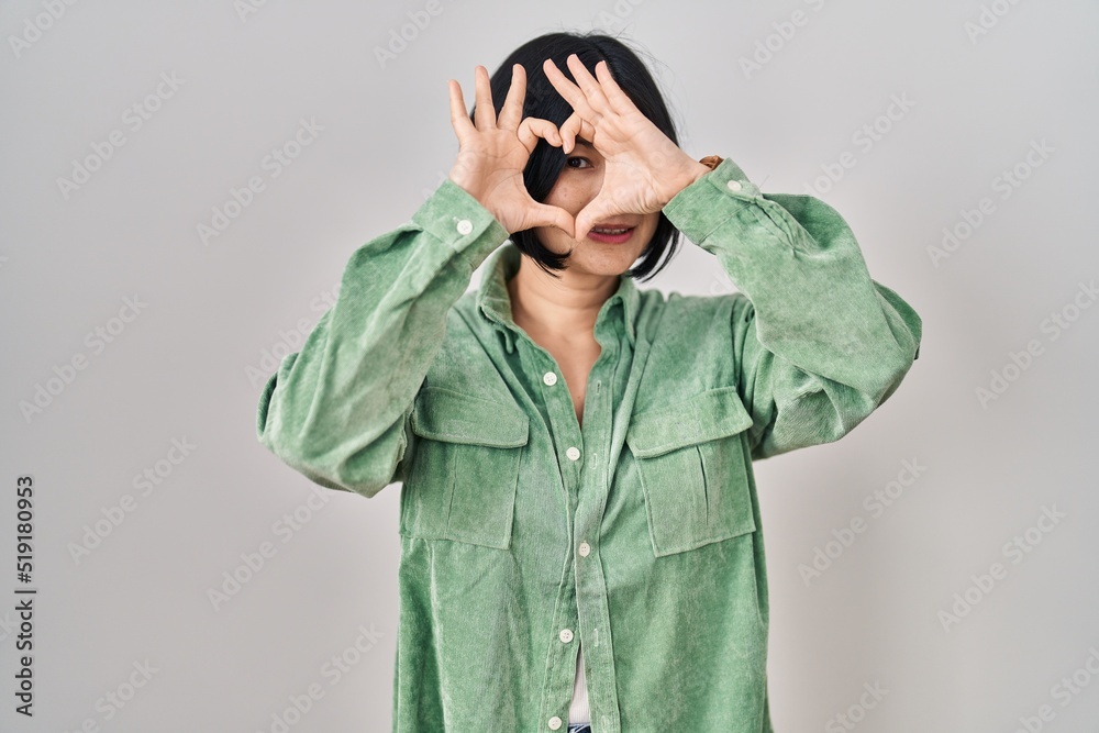 Young asian woman standing over white background doing heart shape with hand and fingers smiling looking through sign Young asian woman standing over white background doing heart shape with hand and fingers smiling looking through sign