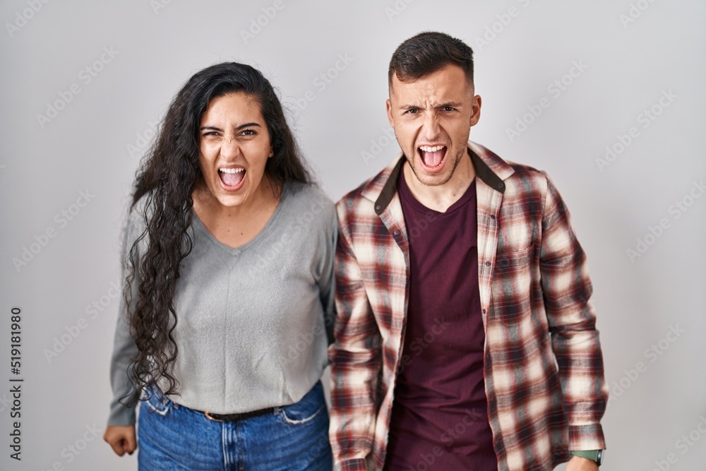 Fototapeta premium Young hispanic couple standing over white background angry and mad screaming frustrated and furious, shouting with anger. rage and aggressive concept.