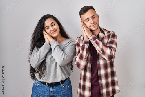 Young hispanic couple standing over white background sleeping tired dreaming and posing with hands together while smiling with closed eyes.