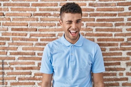 Brazilian young man standing over brick wall winking looking at the camera with sexy expression, cheerful and happy face.