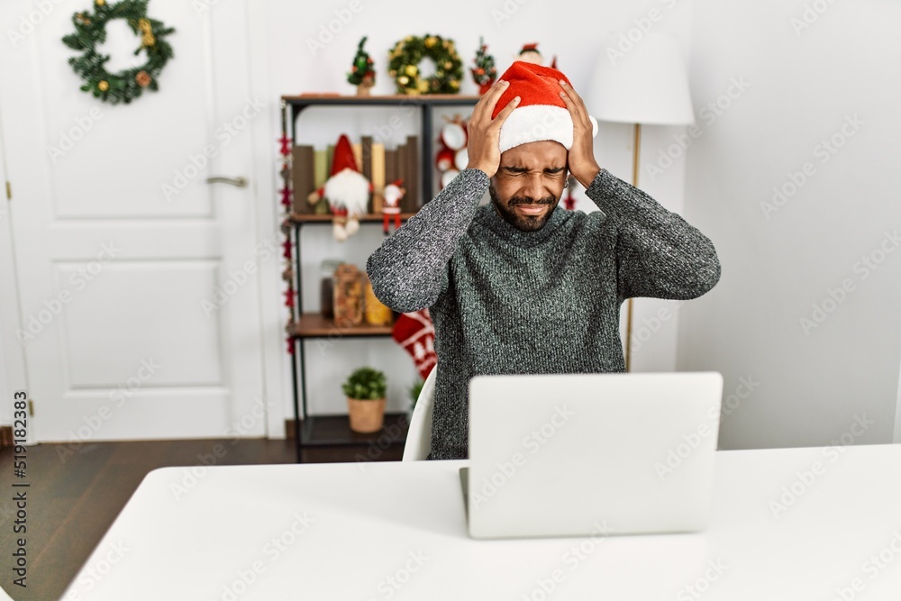 Young hispanic man with beard wearing christmas hat using laptop suffering from headache desperate and stressed because pain and migraine. hands on head. Young hispanic man with beard wearing christmas hat using laptop suffering from headache desperate and stressed because pain and migraine. hands on head.