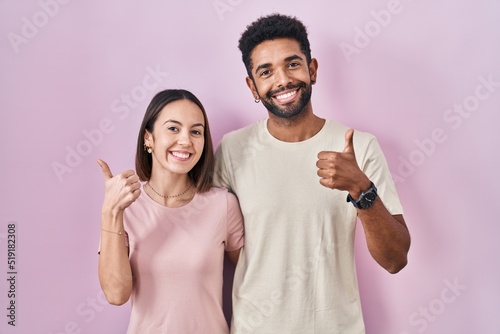 Young hispanic couple together over pink background doing happy thumbs up gesture with hand. approving expression looking at the camera showing success.