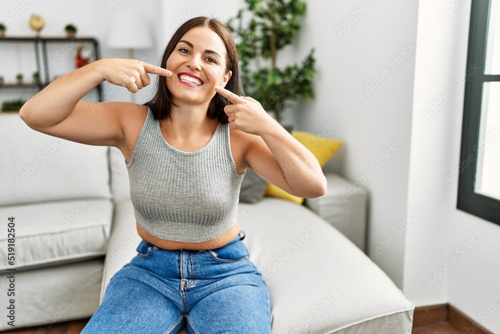 Young brunette woman sitting on the sofa at home smiling cheerful showing and pointing with fingers teeth and mouth. dental health concept. Young brunette woman sitting on the sofa at home smiling cheerful showing and pointing with fingers teeth and mouth. dental health concept.