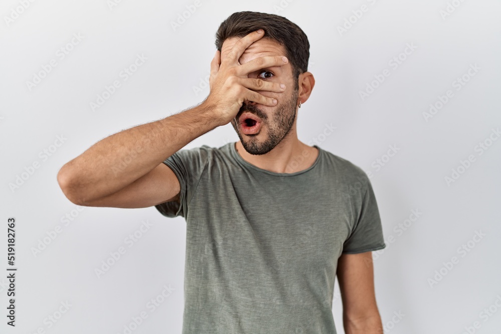 Young hispanic man with beard wearing casual t shirt over white background peeking in shock covering face and eyes with hand, looking through fingers with embarrassed expression. Young hispanic man with beard wearing casual t shirt over white background peeking in shock covering face and eyes with hand, looking through fingers with embarrassed expression.