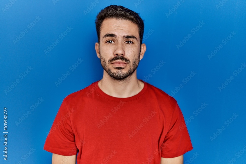 Obraz premium Young hispanic man with beard wearing red t shirt over blue background relaxed with serious expression on face. simple and natural looking at the camera.