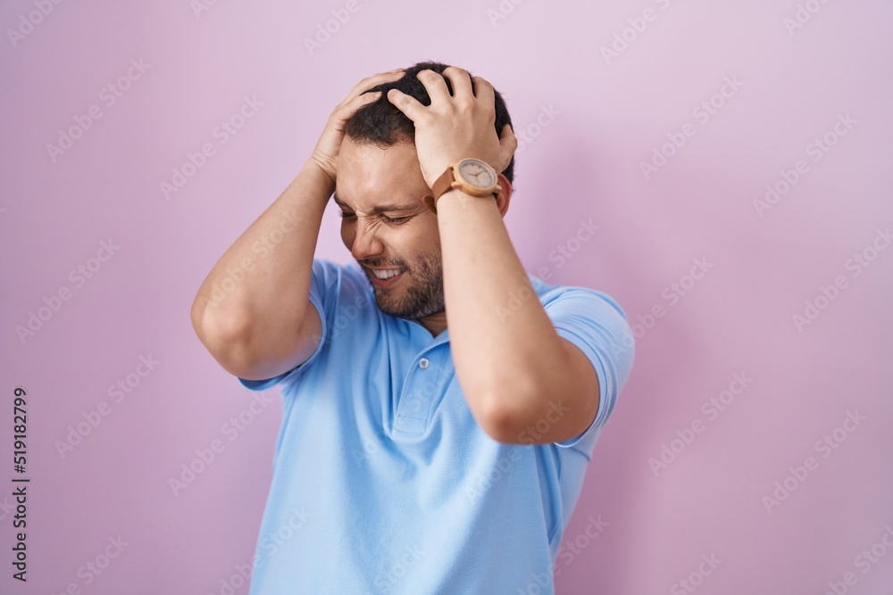 Hispanic man standing over pink background suffering from headache desperate and stressed because pain and migraine. hands on head.