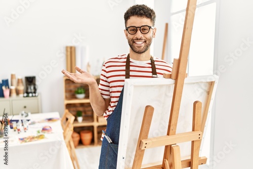Arab young man at art studio pointing aside with hands open palms showing copy space, presenting advertisement smiling excited happy
