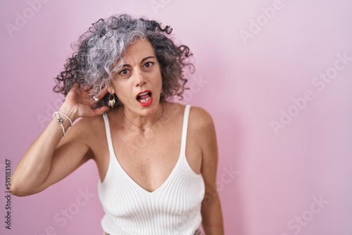 Middle age woman with grey hair standing over pink background smiling with hand over ear listening an hearing to rumor or gossip. deafness concept.