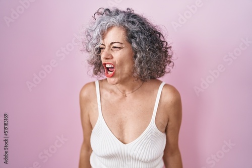 Middle age woman with grey hair standing over pink background angry and mad screaming frustrated and furious, shouting with anger. rage and aggressive concept.