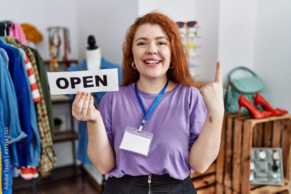 Young redhead woman holding banner with open text at retail shop smiling with an idea or question pointing finger with happy face, number one Young redhead woman holding banner with open text at retail shop smiling with an idea or question pointing finger with happy face, number one