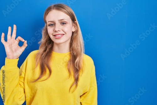Young caucasian woman standing over blue background smiling positive doing ok sign with hand and fingers. successful expression.