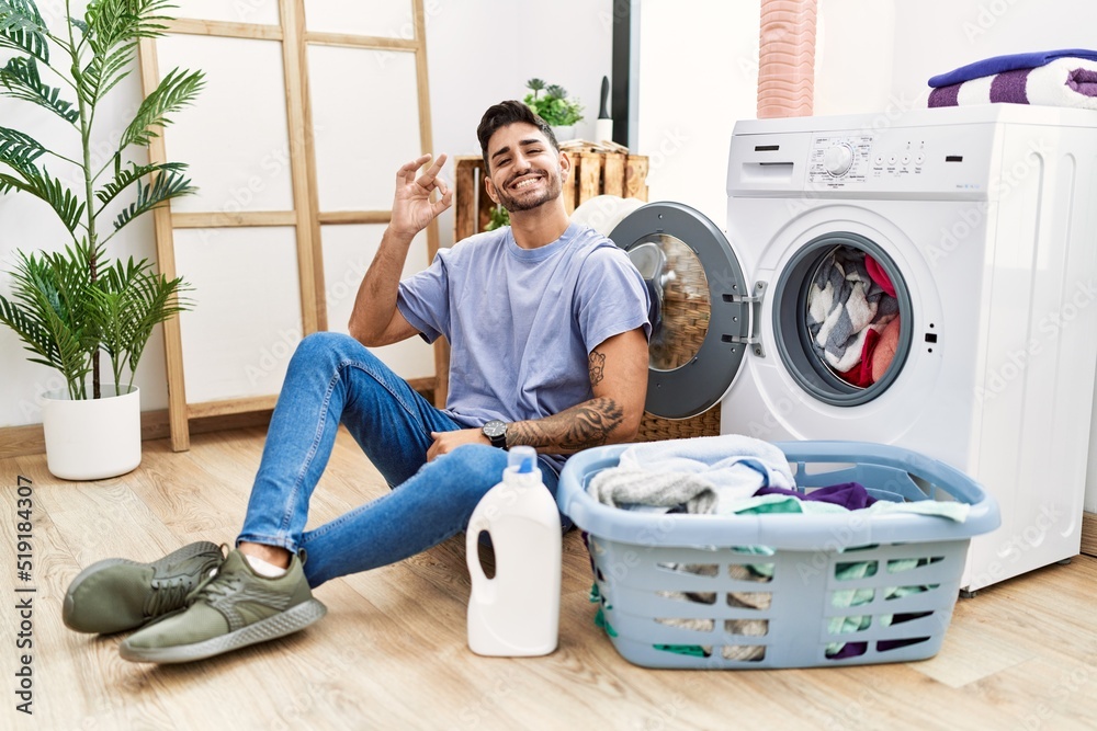 Young hispanic man putting dirty laundry into washing machine smiling positive doing ok sign with hand and fingers. successful expression. Young hispanic man putting dirty laundry into washing machine smiling positive doing ok sign with hand and fingers. successful expression.