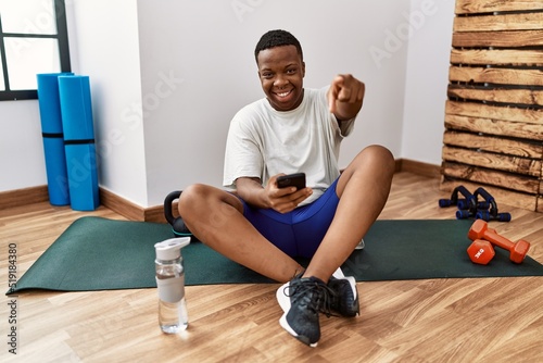 Young african man sitting on training mat at the gym using smartphone pointing to you and the camera with fingers, smiling positive and cheerful