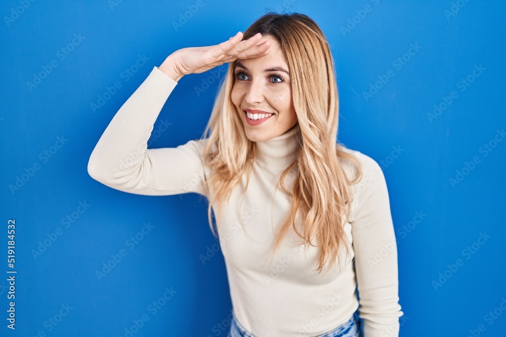 Young caucasian woman standing over blue background very happy and smiling looking far away with hand over head. searching concept. Young caucasian woman standing over blue background very happy and smiling looking far away with hand over head. searching concept.