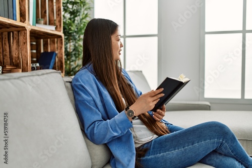Young chinese girl reading book sitting on the sofa at home.
