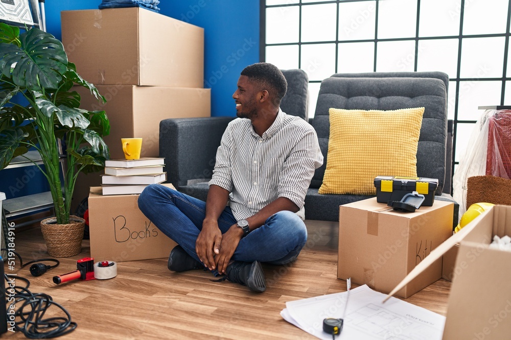 African american man sitting on the floor at new home looking to side, relax profile pose with natural face and confident smile.